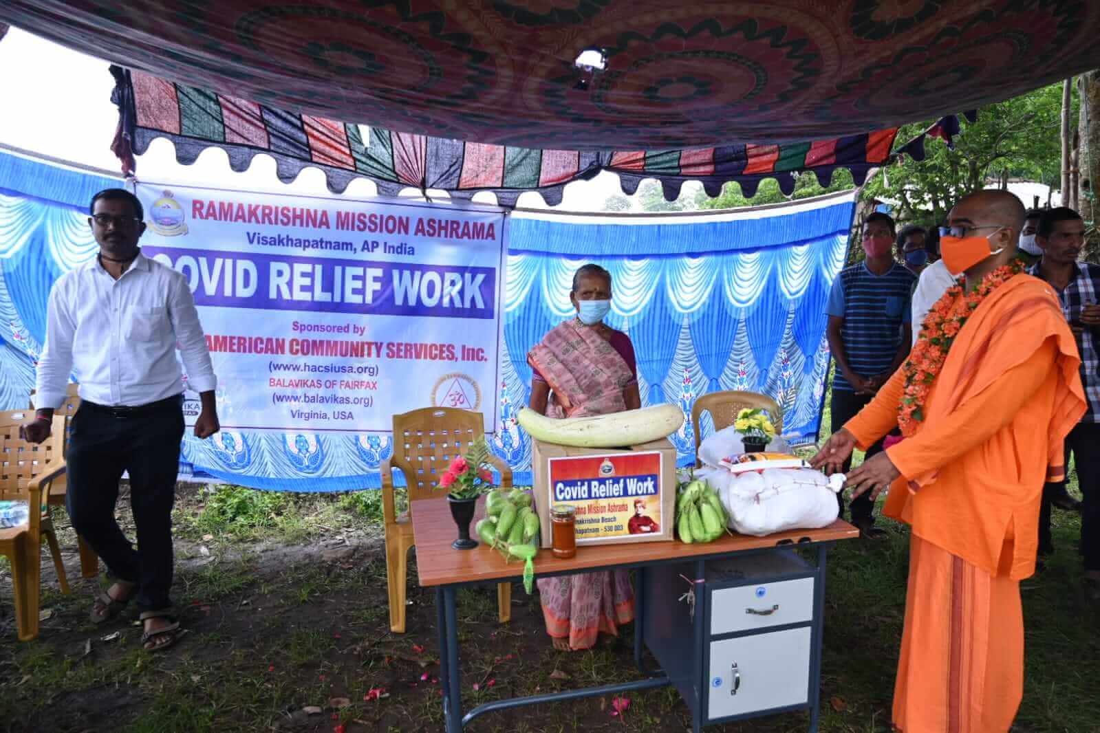 Two people stand behind a table with food and relief supplies under a decorated tent.