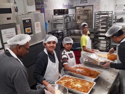 Children and adults preparing food in a kitchen wearing hairnets and aprons.