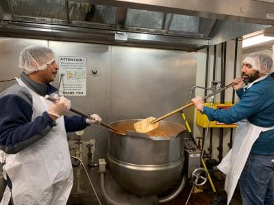 Two people stirring a large pot in an industrial kitchen.